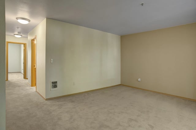 Empty living room with neutral walls and beige carpet, leading to a hallway with two doors.