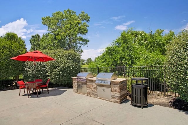Outdoor grilling station with two built-in grills, a stone counter, and a trash can, next to a red umbrella and table.