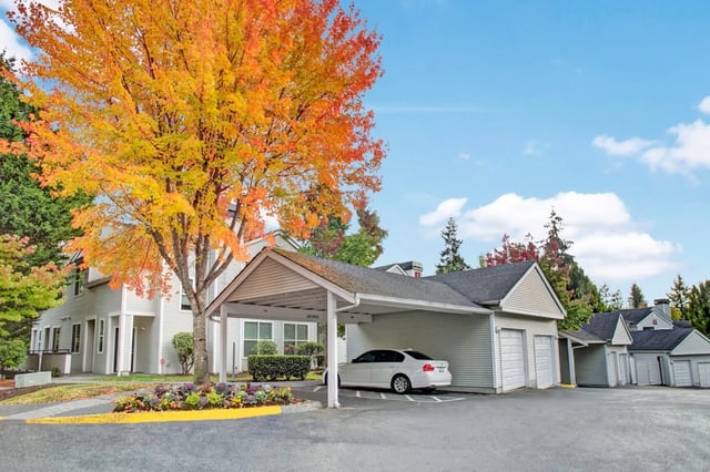 Apartment building exterior with a covered parking spot and a white car, surrounded by colorful autumn trees.