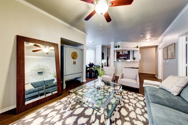 Living room in an apartment with a glass coffee table, large mirror, and open kitchen visible in the background.