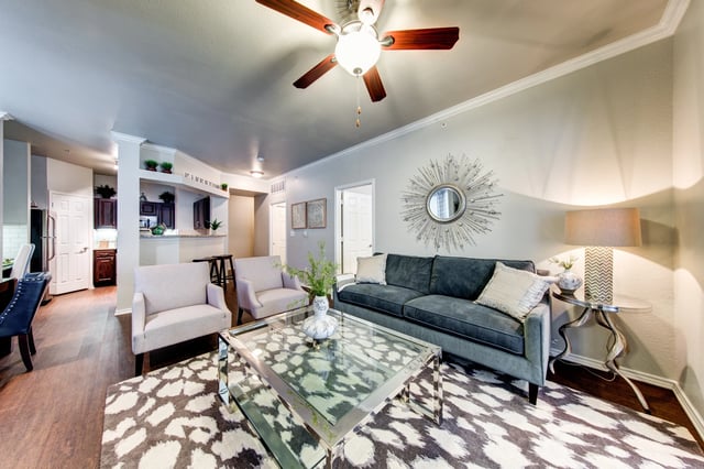 Living room in an apartment with a gray sofa, two chairs, glass coffee table, rug, and ceiling fan.