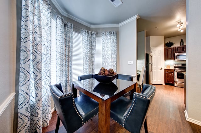 Dining area with glass-top table and blue upholstered chairs; patterned curtains and open kitchen.