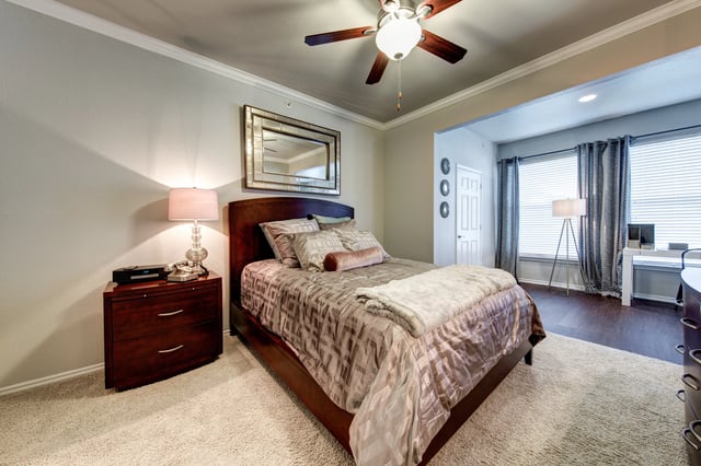 Bedroom in an apartment with a dark wood bed, nightstand, lamp, ceiling fan, and window with curtains.
