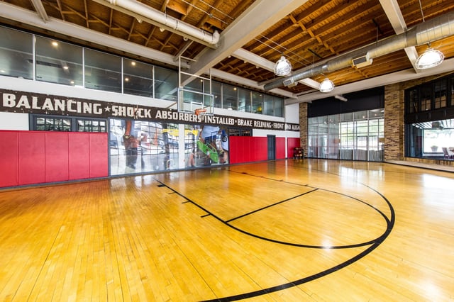 Indoor basketball court with wooden floor, hoop, padded walls, and exposed ceiling beams.