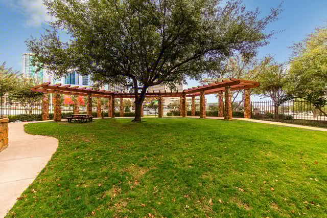 Park-like green space with a pergola, stone columns, and a picnic table under trees.