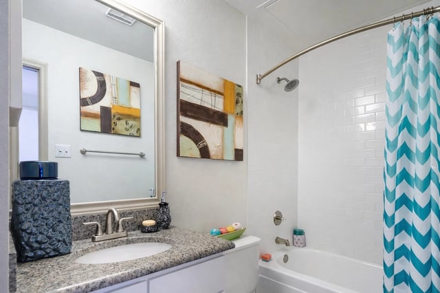 Modern bathroom with a granite countertop vanity, sink, mirror, and a white tiled shower with a teal chevron curtain.