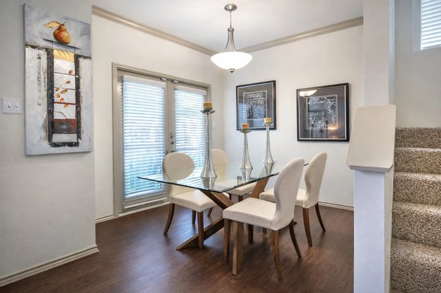 A dining room with a glass-top table and four chairs, featuring a painting and a staircase.