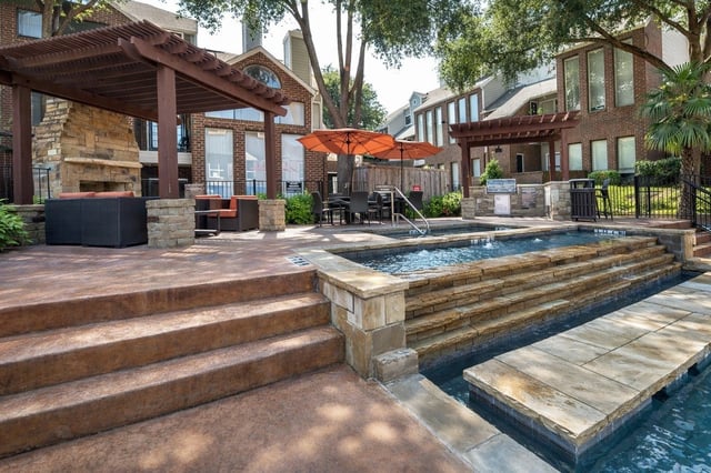 Outdoor swimming pool and spa area with lounge seating and a fireplace under a pergola.