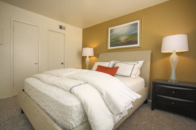 A neatly made bed with white linens and a burnt orange accent pillow, next to a nightstand with a lamp.