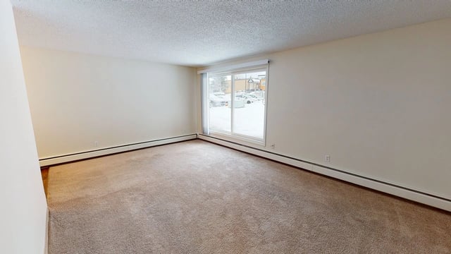 Empty apartment living room with carpet and a large sliding glass door showing a snowy parking lot.