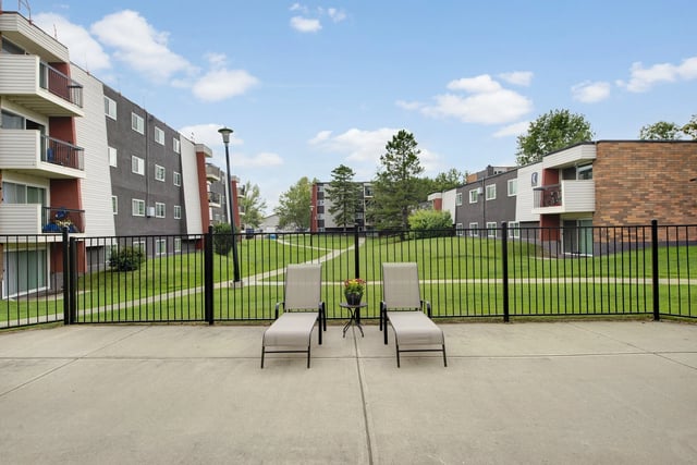 Two lounge chairs and a small table on a patio overlooking a grassy courtyard and apartment buildings.