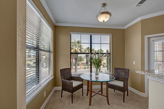 Dining area with glass-top table and two chairs, large windows with blinds overlooking palm trees.