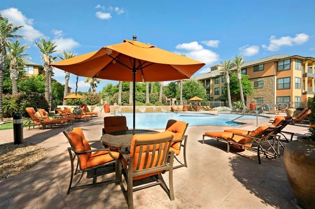 Outdoor swimming pool with lounge chairs and umbrellas, surrounded by palm trees and apartment buildings.