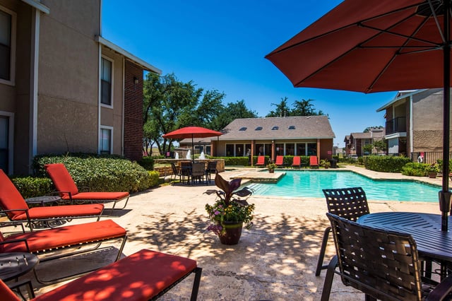 Outdoor apartment community pool area with red lounge chairs, umbrellas, and surrounding buildings.