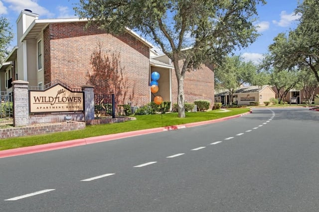 Exterior view of Wildflower Apartments entry with brick building, sign, and landscaping along a curved road.
