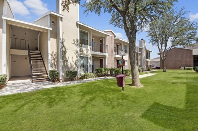Exterior view of a multi-family apartment complex with green lawn, trees, and walkways.