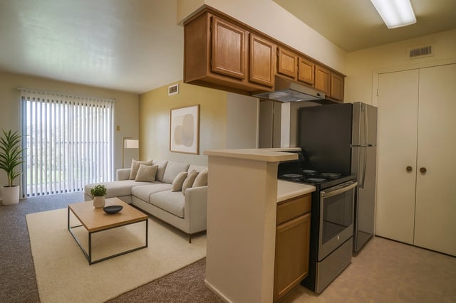Open-concept living room with beige sofa, coffee table, plant, and a kitchen with stainless steel appliances.