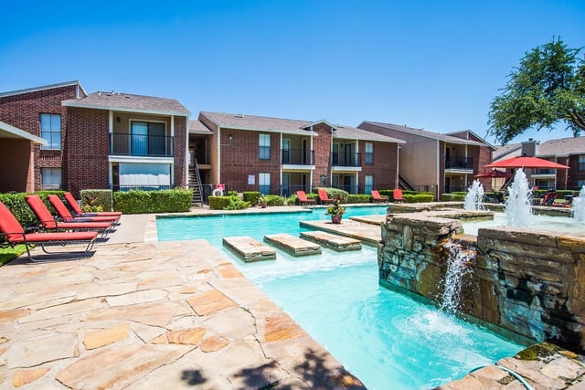 Outdoor pool area with stone deck, red lounge chairs, and brick apartment buildings in the background.
