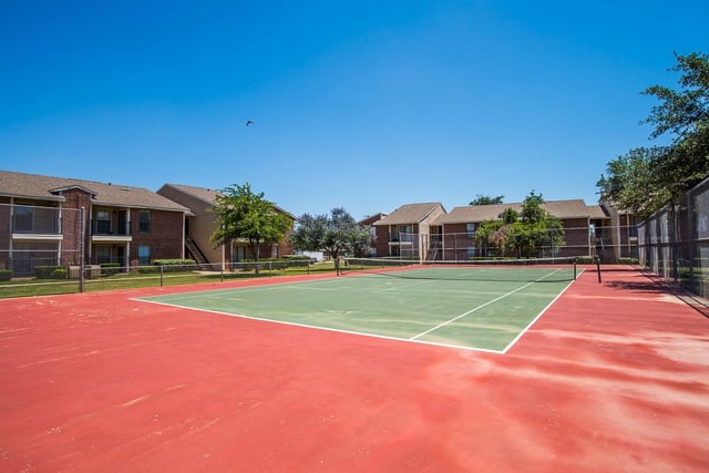 Outdoor tennis court at a multifamily community, with brick buildings and a chain-link fence.