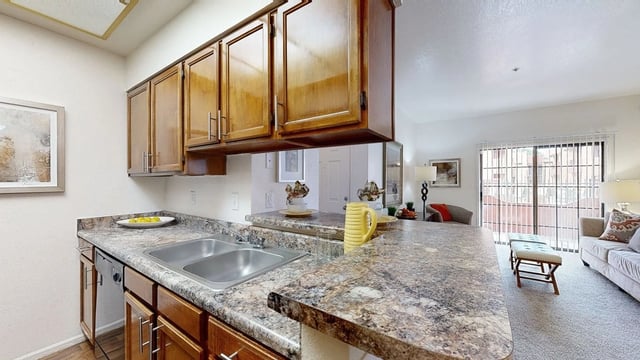 Kitchen with granite countertops, wooden cabinets, and stainless steel sink. Living area visible in background.