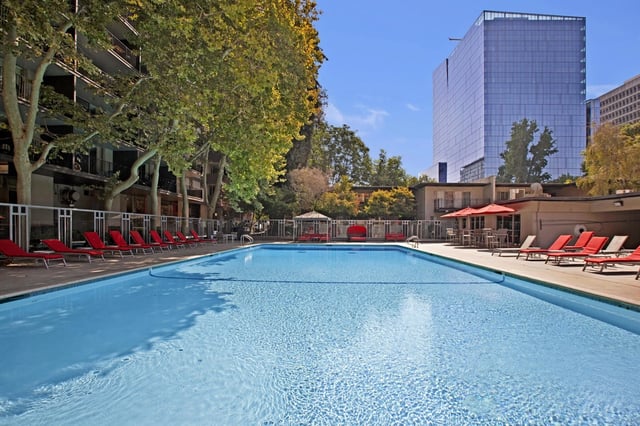 Swimming pool with lounge chairs and umbrellas, surrounded by trees and buildings.