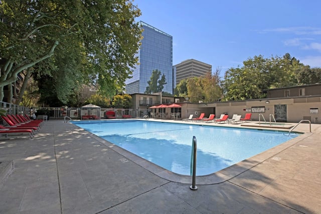 Swimming pool with lounge chairs and umbrellas, surrounded by trees and buildings.