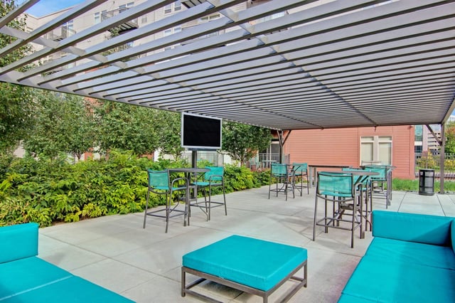 Outdoor community lounge with turquoise seating, high-top tables, planters, and a wall-mounted TV under a metal pergola.