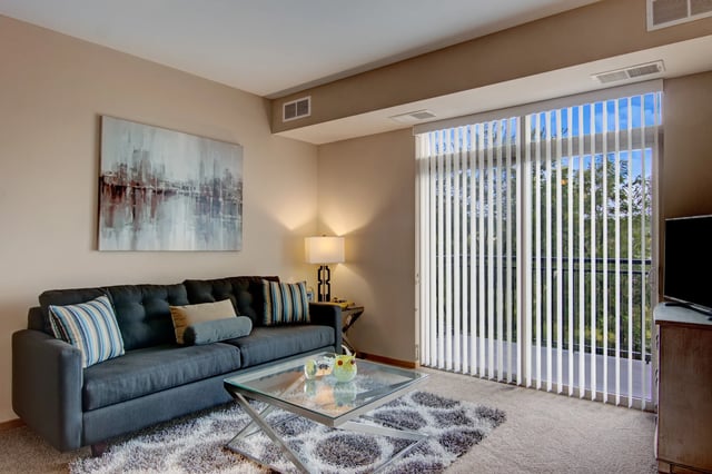 Apartment living room with a gray sofa, glass coffee table, and sliding doors with vertical blinds.