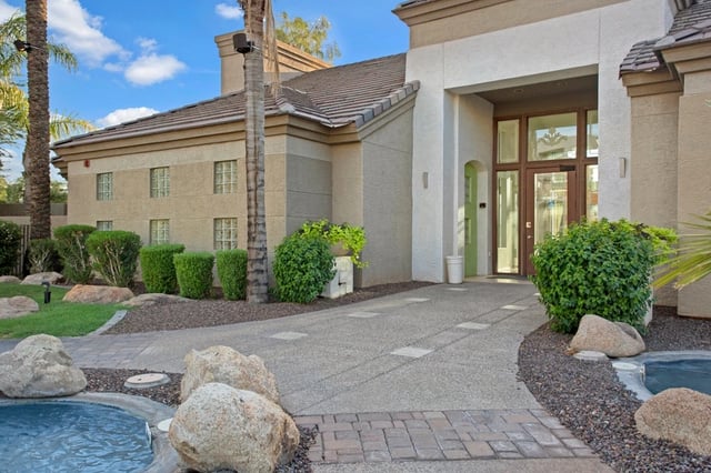 Exterior of apartment building entrance with a walkway, landscaping, and a pool.