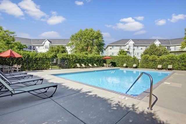 Outdoor swimming pool with lounge chairs and umbrellas, surrounded by greenery and apartment buildings.