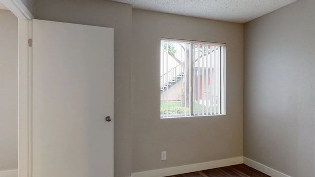 Interior view of an empty apartment room with a closed door and a window showing stairs outside.