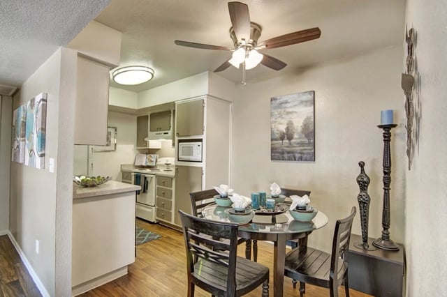Dining area with a round glass-top table and four chairs, adjacent to a kitchen with white appliances.