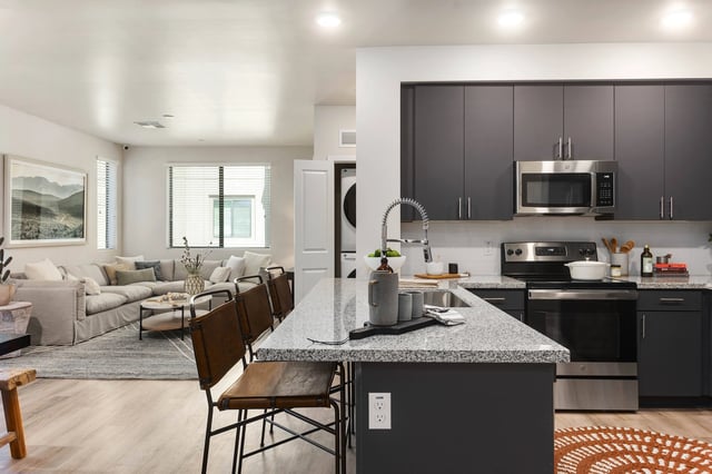 Modern kitchen with granite island and stainless steel appliances, open to a living room with a sectional sofa.