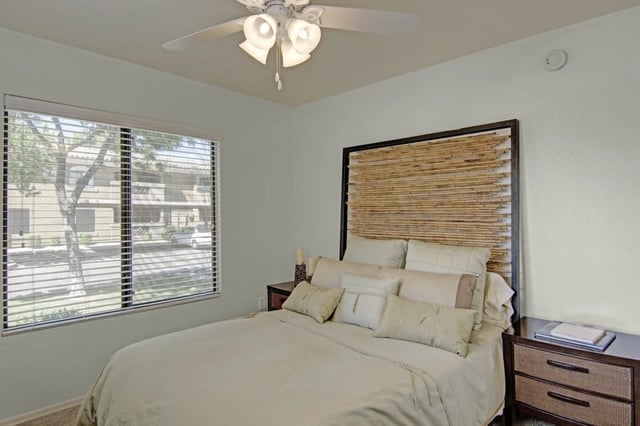 Queen bed with beige linens and a bamboo headboard in a bedroom with blinds and a ceiling fan.