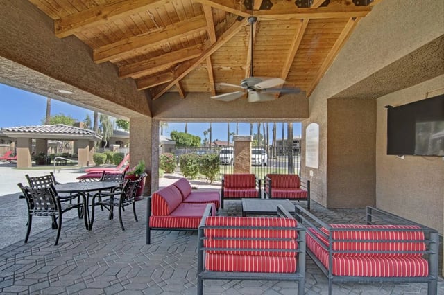 Covered outdoor seating area with red and white striped cushions and a ceiling fan, overlooking a pool area.