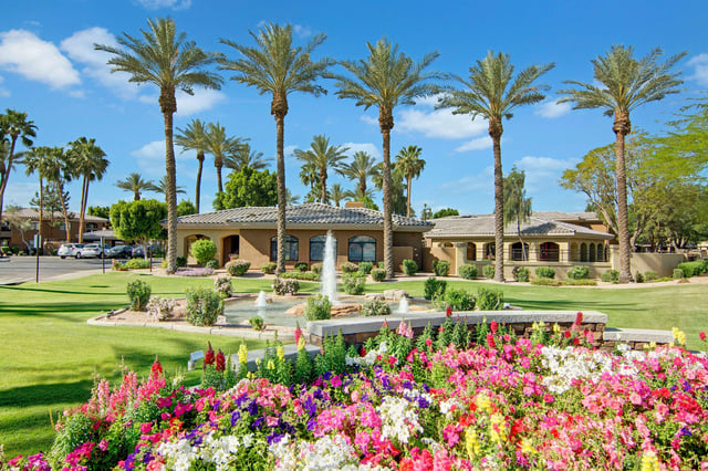 Manicured lawn with a fountain and palm trees outside a tan stucco building, with a flower bed in the foreground.