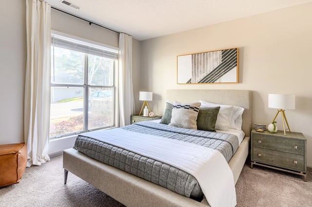 A staged bedroom with a large window, bed, and nightstand.