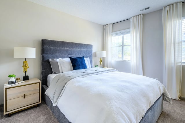 Comfortable king-sized bed with grey headboard, white linens, and decorative pillows. Nightstand with lamp on the left.