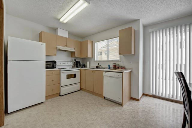 Kitchen with white appliances, light wood cabinets, and a window with blinds.