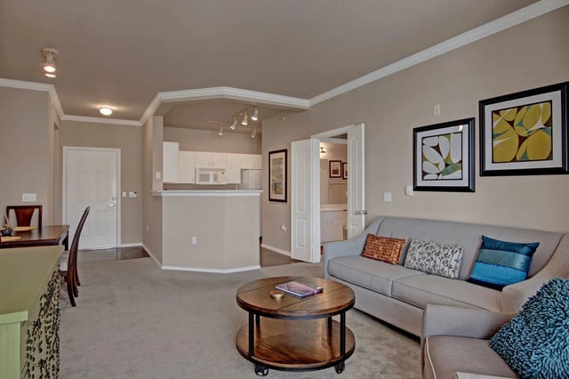 Living room with sofa, coffee table, and dining area, with kitchen visible in the background.