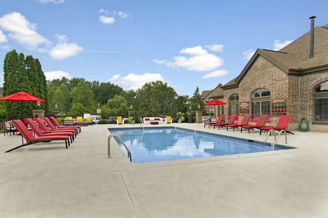 Outdoor swimming pool with red lounge chairs and umbrellas, and a brick building in the background.