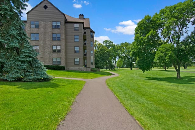 Apartment building exterior with a paved walkway through manicured lawns and mature trees under a blue sky.