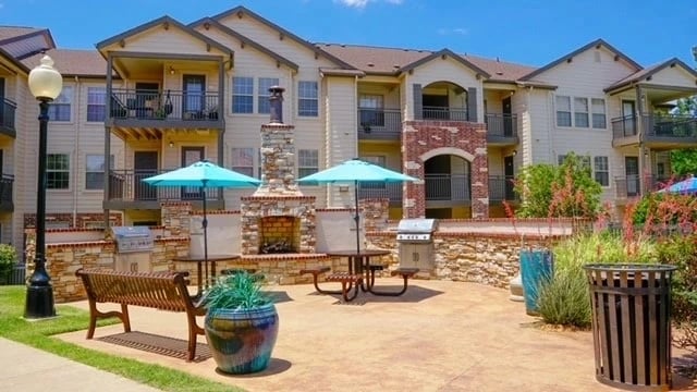 Outdoor community courtyard with a stone fireplace, benches, and umbrellas in front of apartment buildings.