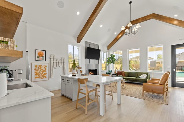 Open-concept living area with vaulted white ceiling, exposed wooden beams, large windows, and a fireplace.
