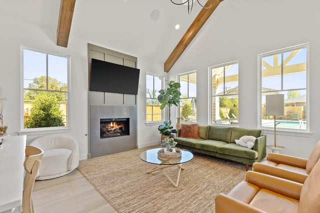 Bright living room with vaulted ceiling, exposed wooden beams, fireplace, wall-mounted TV, and large pool-view windows.