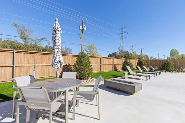 Outdoor community courtyard with a round table, chairs, umbrella, lounge chairs, and a wooden fence.