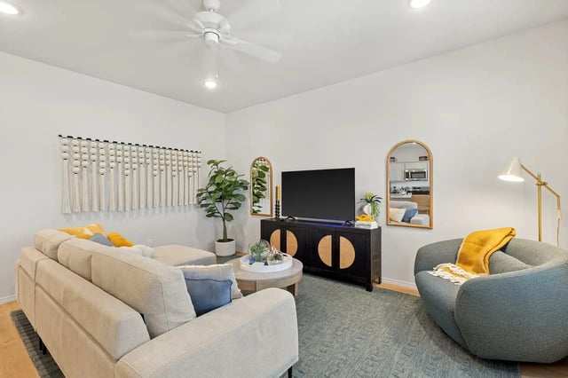 Bright apartment living room with a beige sectional, gray curved chair, TV on a dark cabinet, and potted plants.