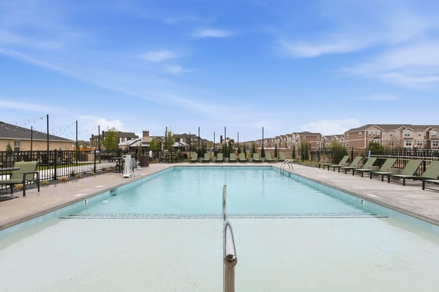 An outdoor swimming pool with lounge chairs and a backdrop of apartment buildings under a blue sky.