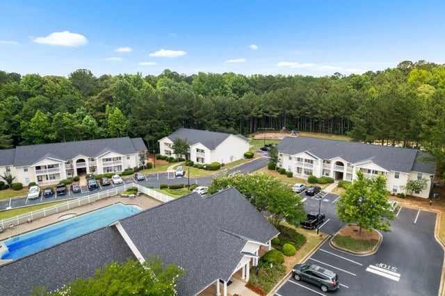 Aerial view of apartment buildings and swimming pool with lush trees and blue sky.