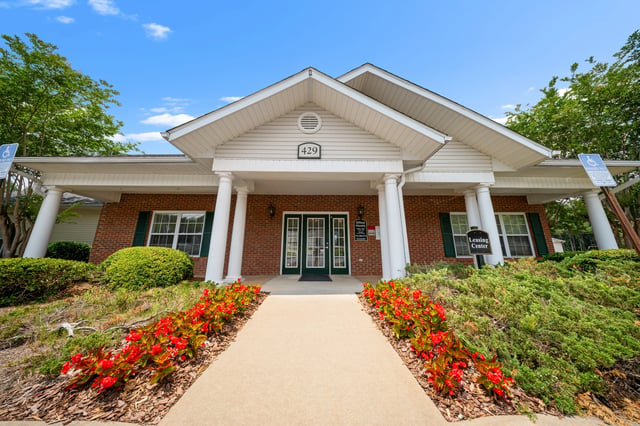 The leasing center building entrance with a paved walkway, flowering plants, and lush greenery at Ashton Pointe apartments in Monroe, GA.